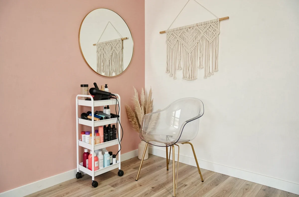 Coin salon de coiffure maison décoré en style bohème avec miroir rond doré, chaise transparente et chariot porte-outils blanc
